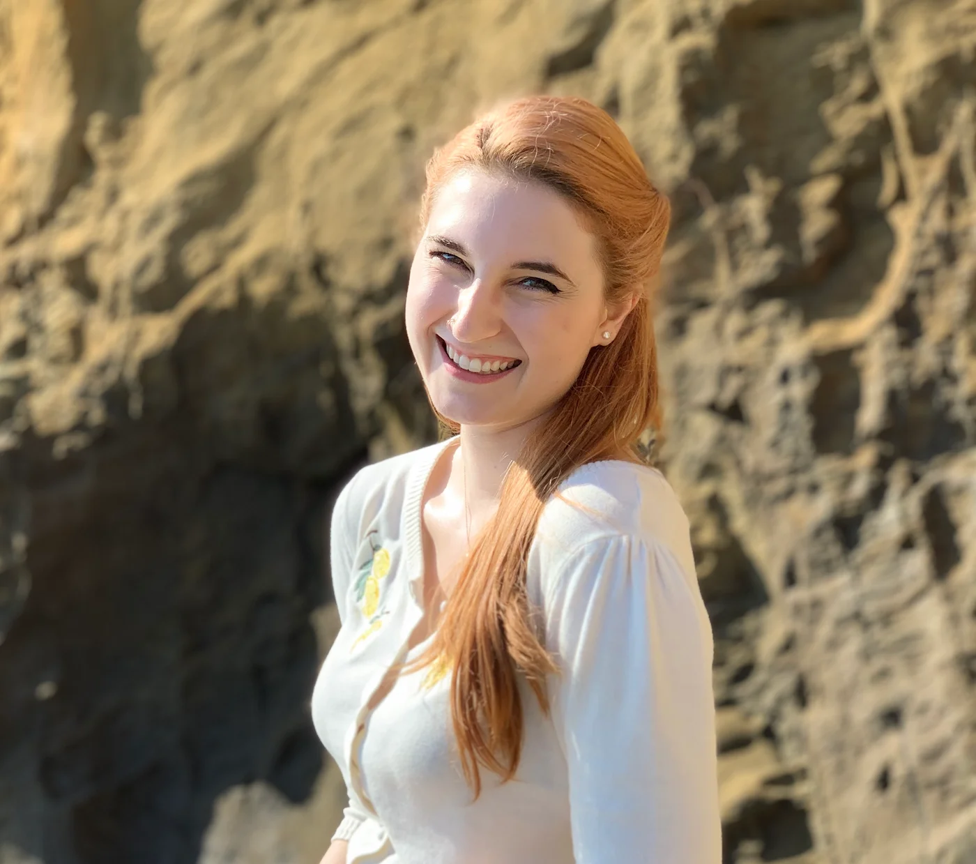 Camille, a white femme with red hair, smiling pictured from the waist up. They're standing outside with large rocks behind them.